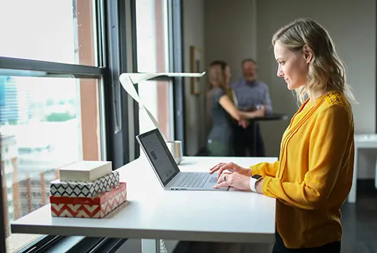 Kvinna i gul tröja står med sin laptop vid ett högt bord