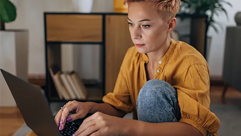 Kvinna i gul skjorta sitter på golvet med sin dator. Hon är väldigt fokuserad.